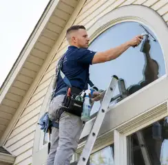Professional window cleaner on a ladder using a squeegee to clean an exterior arched window, Manchester, NH.